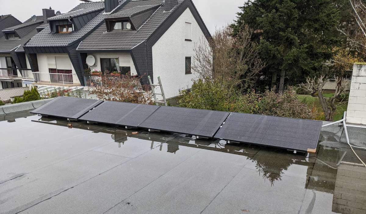 4 Solarpaneele auf einem Flachdach, auf dem gerade etwas Regen steht. Dahinter ein Garten mit Büschen und Bäumen und einige andere Häuser von denen man aber nur die oberen Stockwerke sieht.